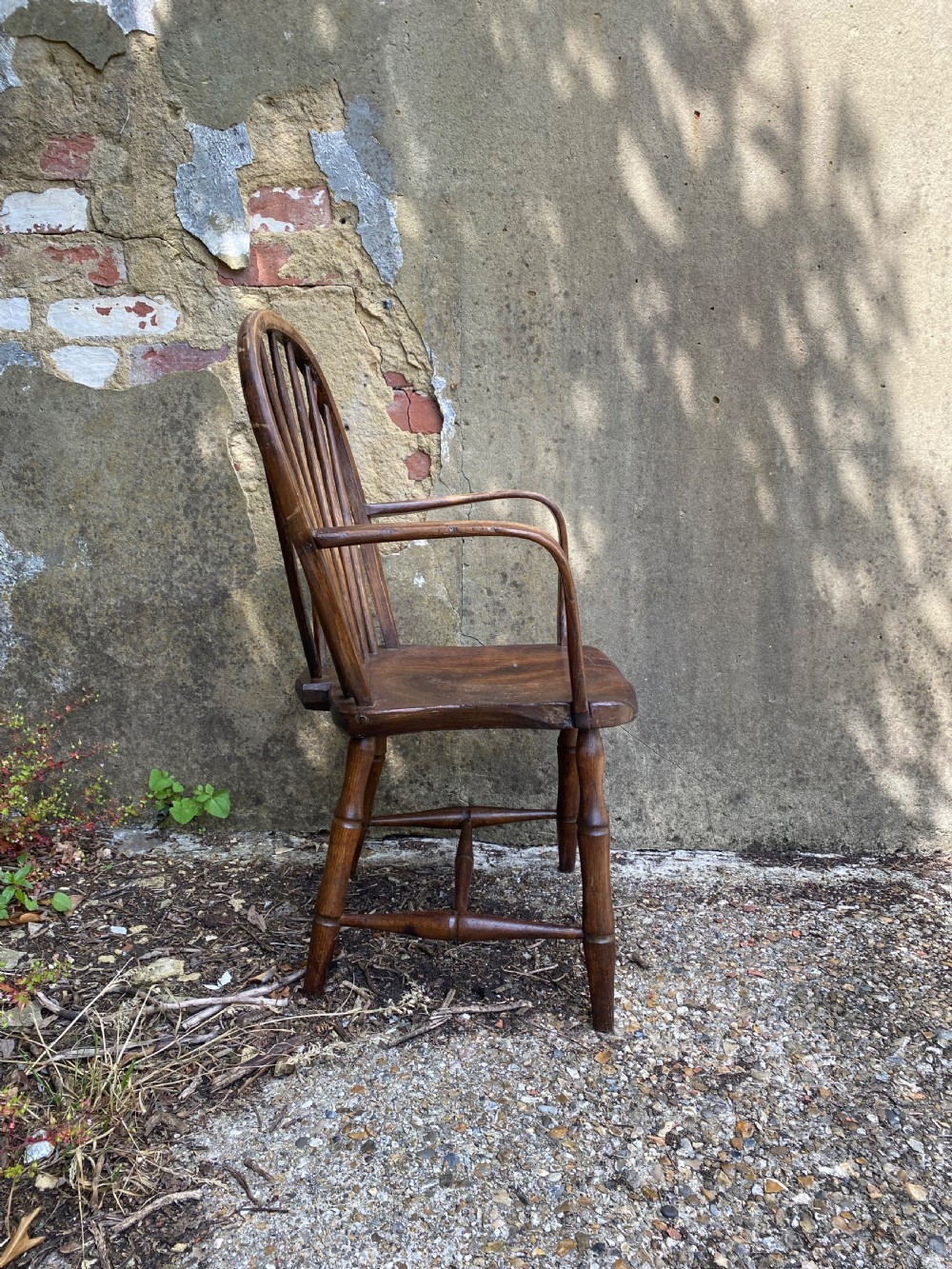 An antique wooden Windsor armchair with a stick-back design and a sculpted wooden seat stands against a weathered wall. View VIII