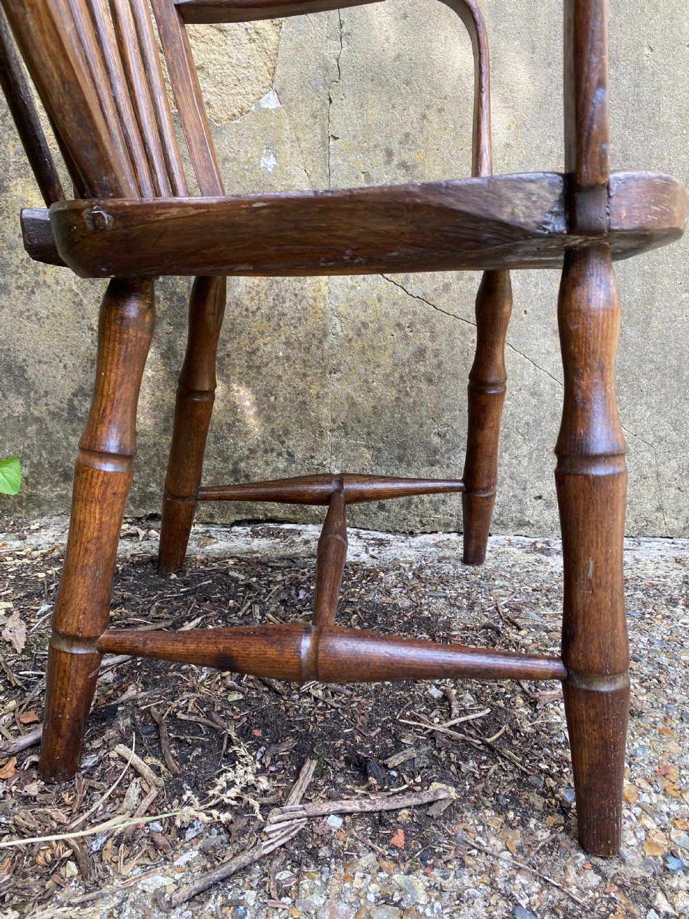 An antique wooden Windsor armchair with a stick-back design and a sculpted wooden seat stands against a weathered wall. View X