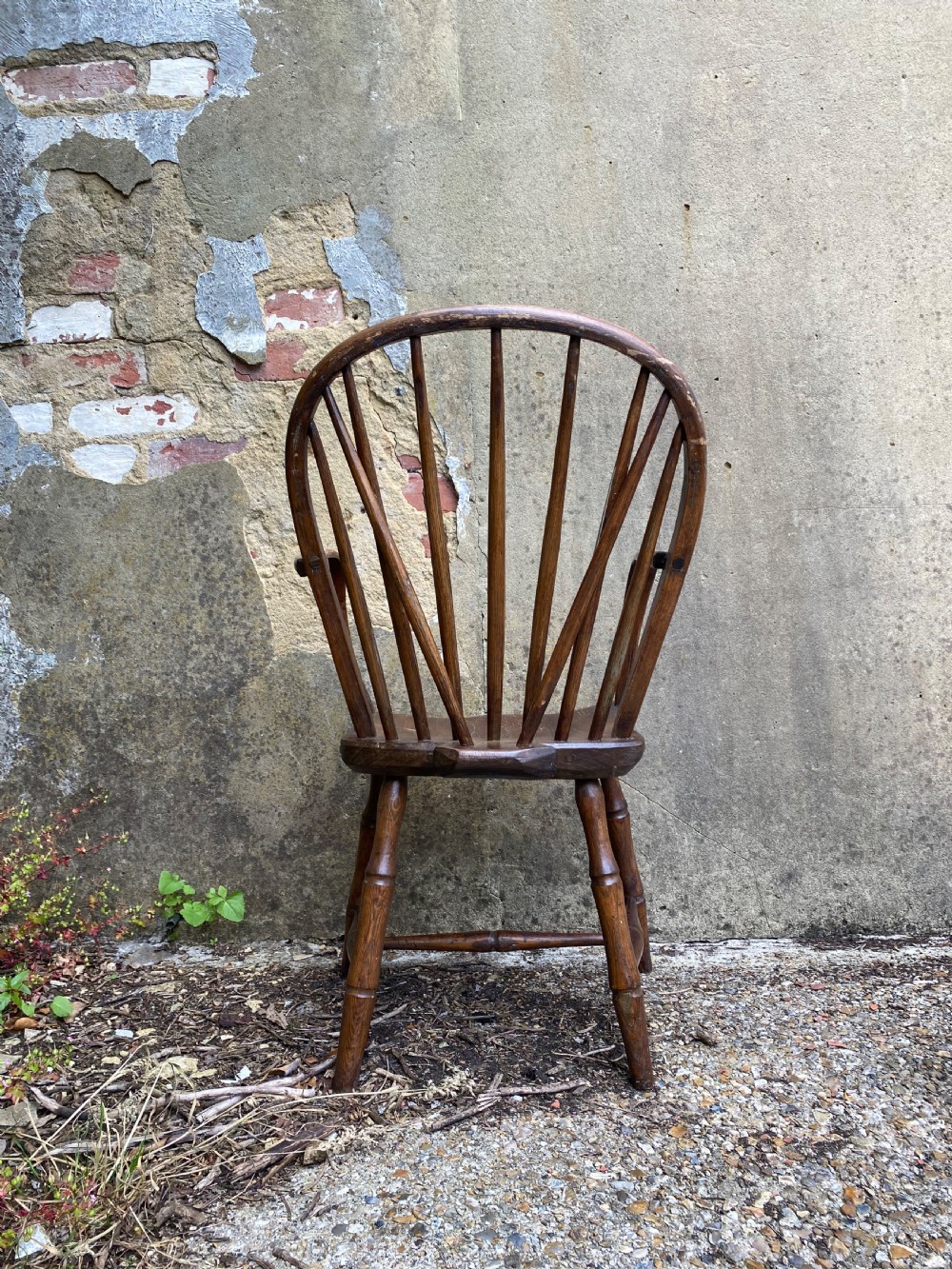 An antique wooden Windsor armchair with a stick-back design and a sculpted wooden seat stands against a weathered wall. View XIV