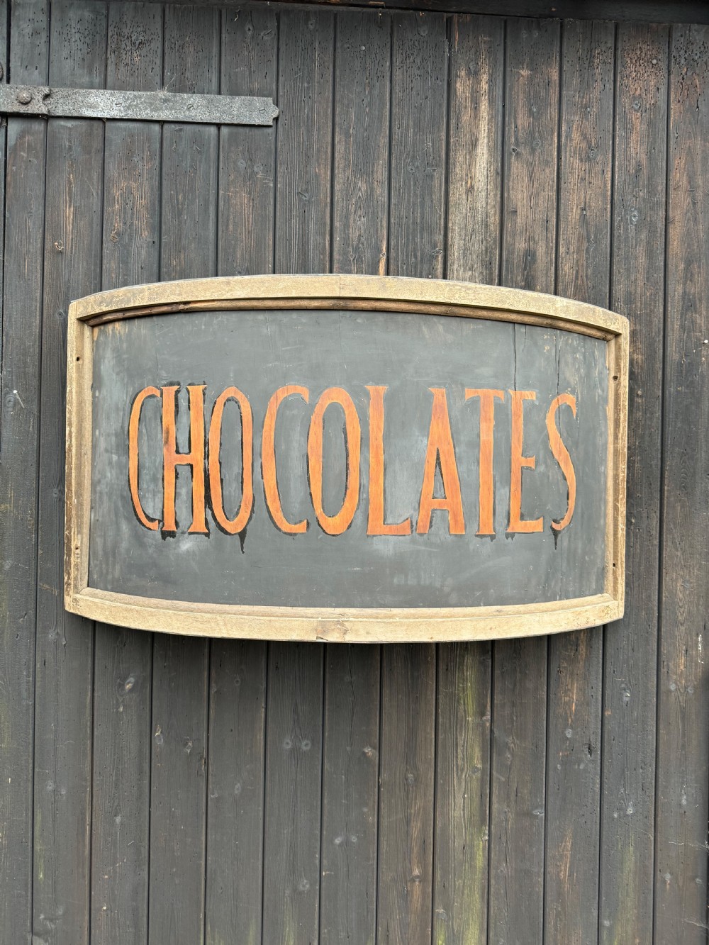 A curved antique wooden trade sign featuring orange hand-painted lettering reading CHOCOLATES on a dark grey background with a natural timber frame.