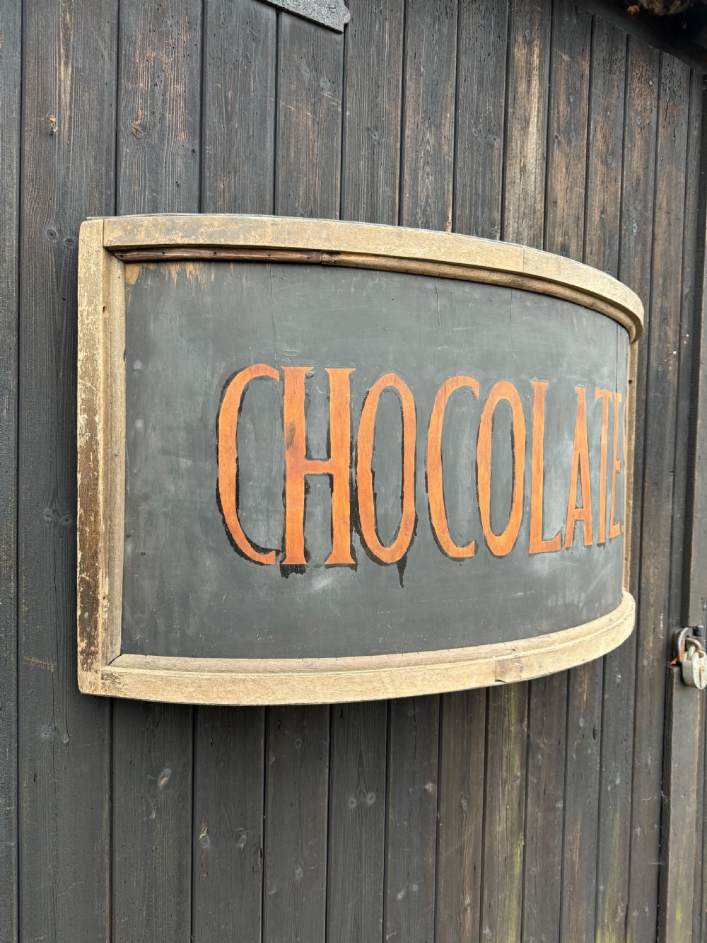 A curved antique wooden trade sign featuring orange hand-painted lettering reading CHOCOLATES on a dark grey background with a natural timber frame. View II