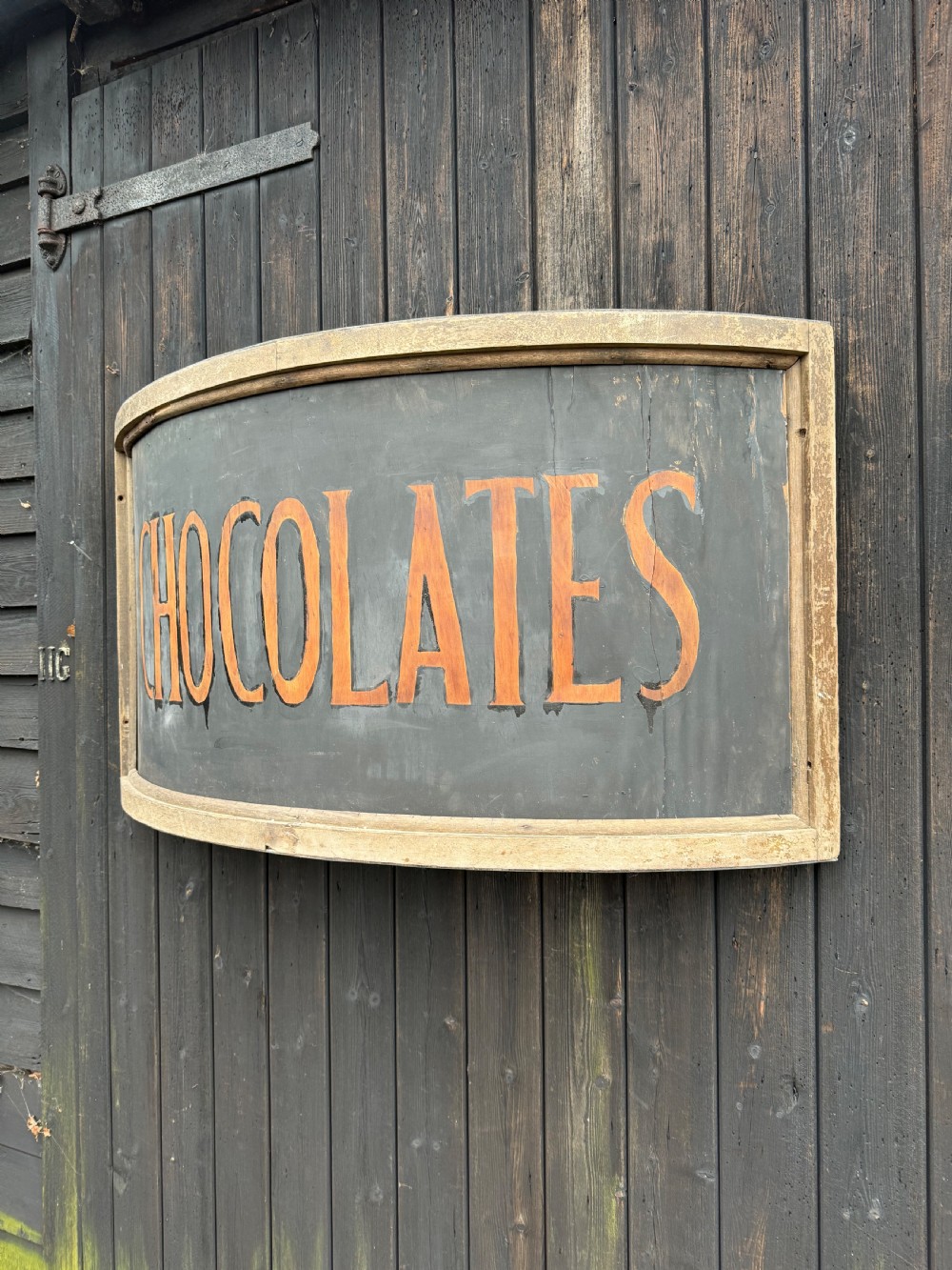 A curved antique wooden trade sign featuring orange hand-painted lettering reading CHOCOLATES on a dark grey background with a natural timber frame. View III
