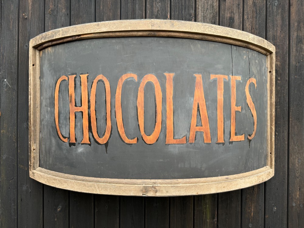 A curved antique wooden trade sign featuring orange hand-painted lettering reading CHOCOLATES on a dark grey background with a natural timber frame. View IV