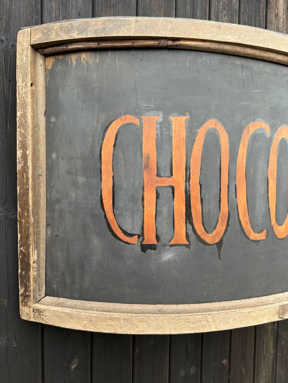 A curved antique wooden trade sign featuring orange hand-painted lettering reading CHOCOLATES on a dark grey background with a natural timber frame. View V