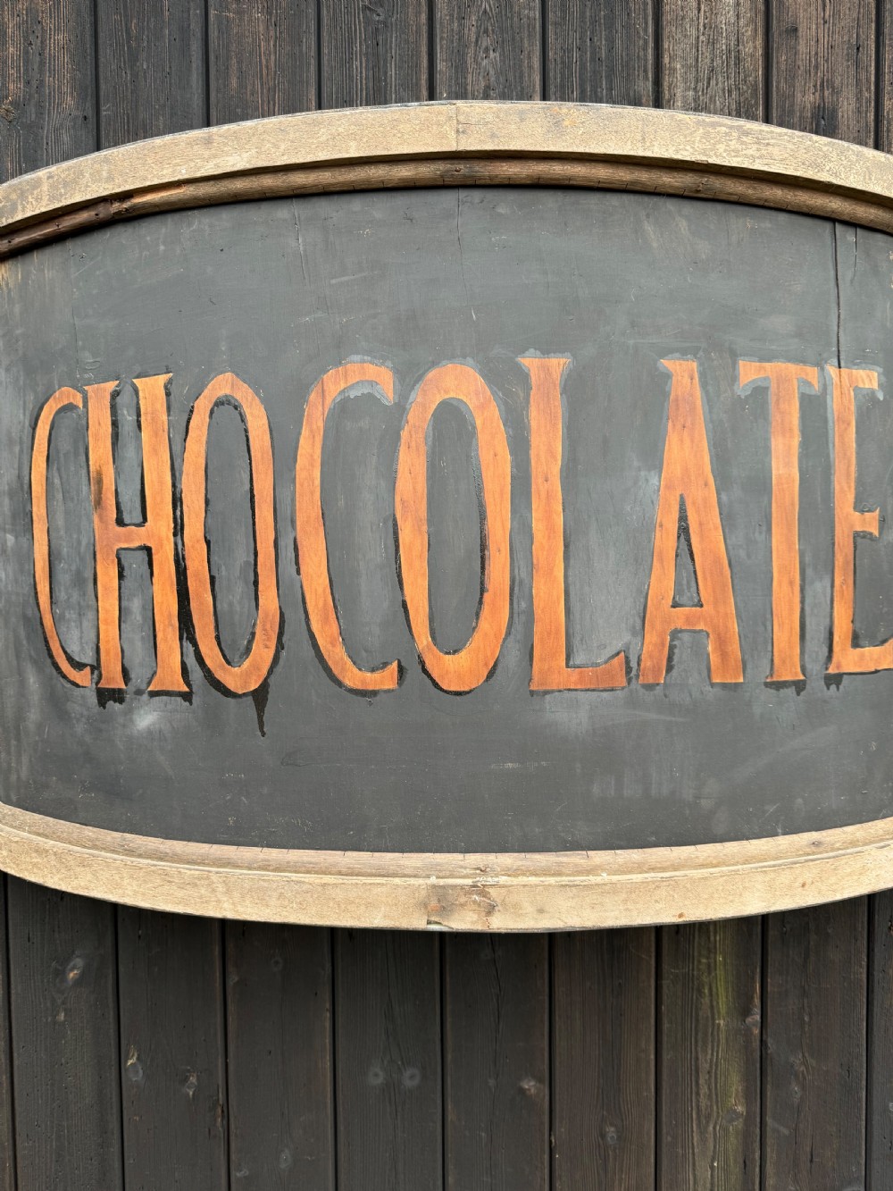 A curved antique wooden trade sign featuring orange hand-painted lettering reading CHOCOLATES on a dark grey background with a natural timber frame. View VI