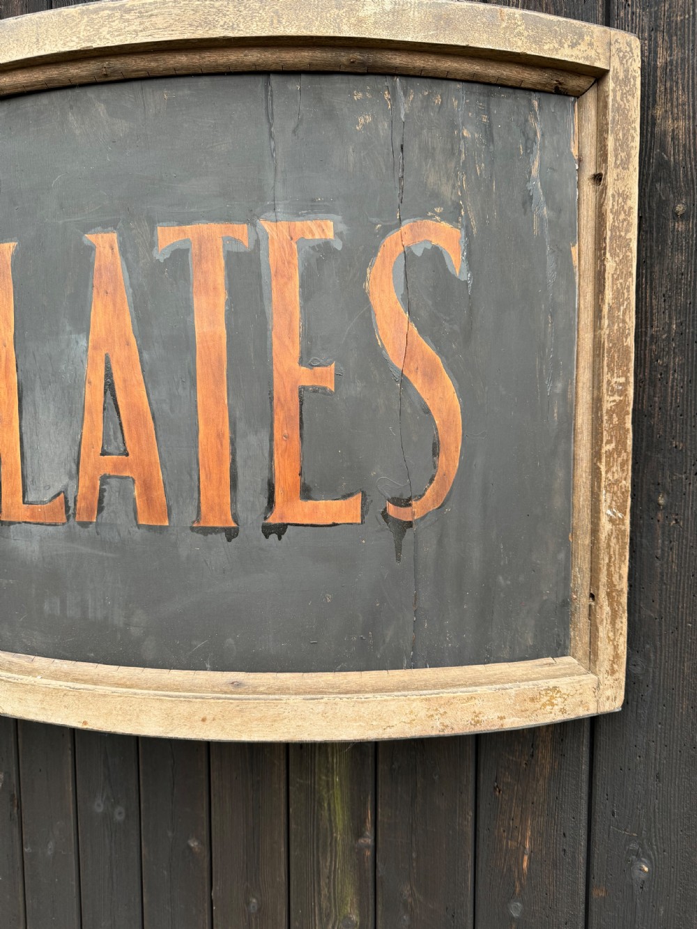 A curved antique wooden trade sign featuring orange hand-painted lettering reading CHOCOLATES on a dark grey background with a natural timber frame. View VII