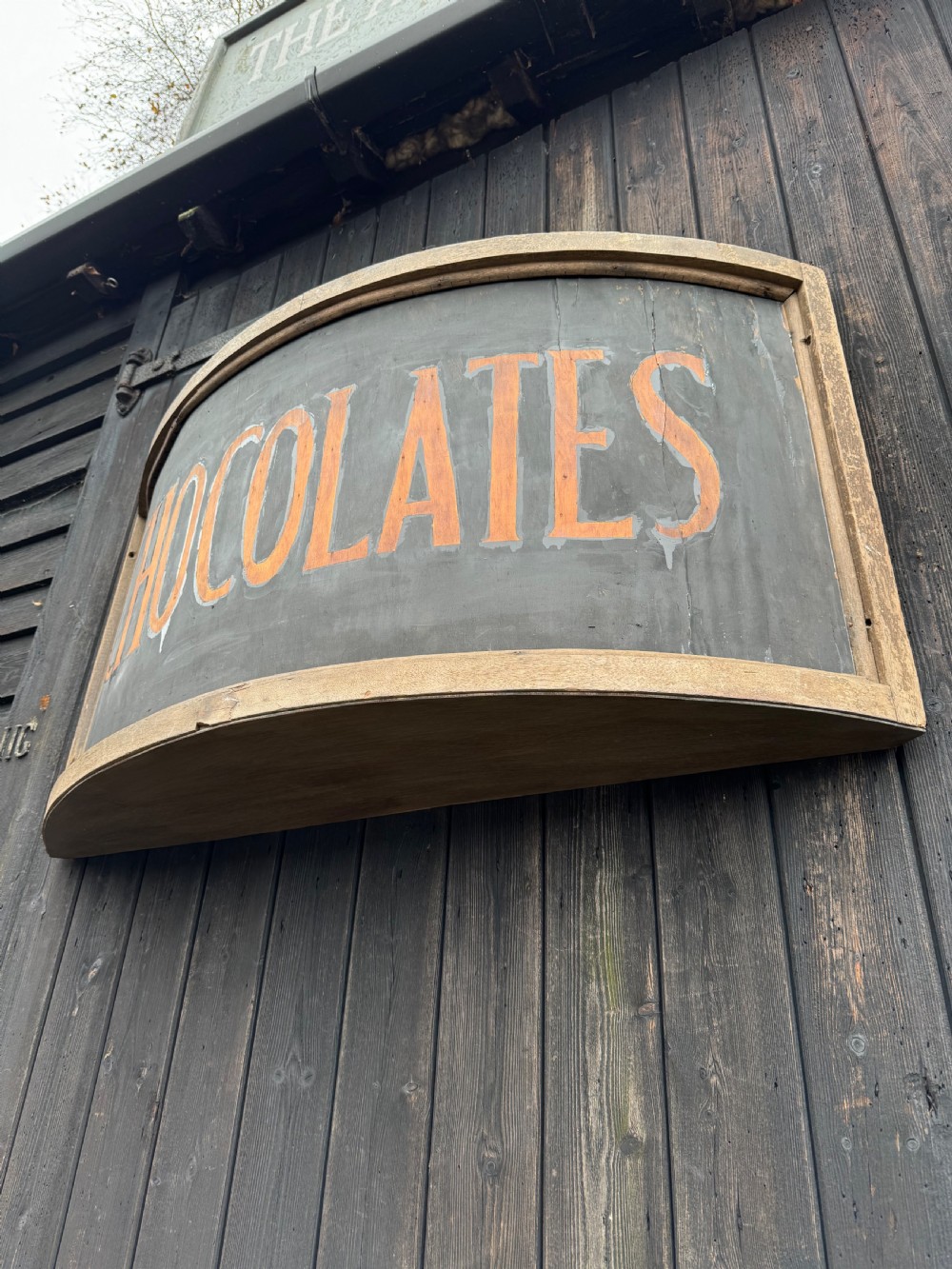 A curved antique wooden trade sign featuring orange hand-painted lettering reading CHOCOLATES on a dark grey background with a natural timber frame. View VIII