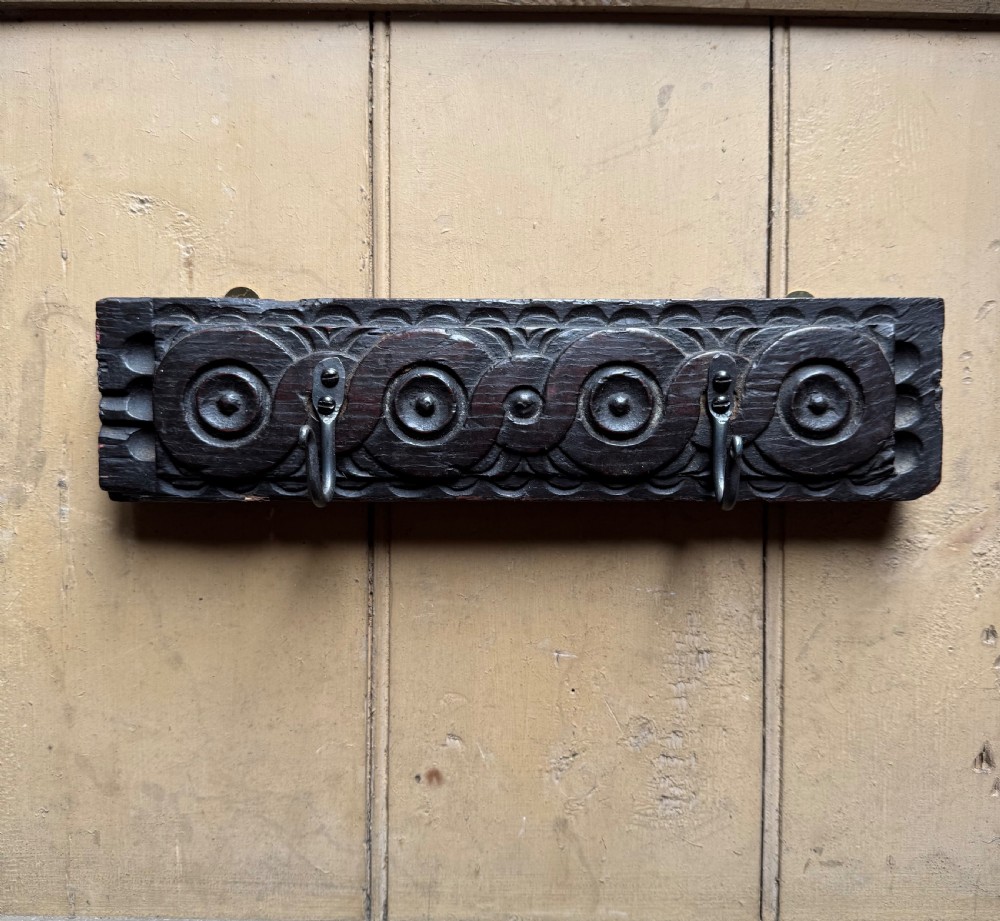 A dark carved oak wooden panel featuring relief carved circular scroll motifs and fitted with two dark metal hooks.