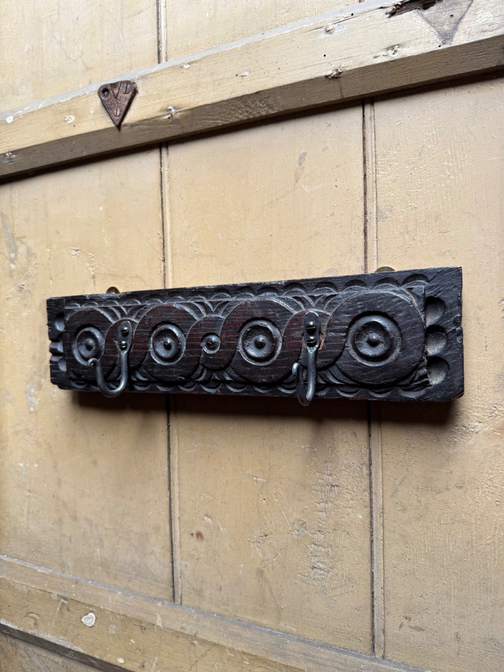 A dark carved oak wooden panel featuring relief carved circular scroll motifs and fitted with two dark metal hooks. View II