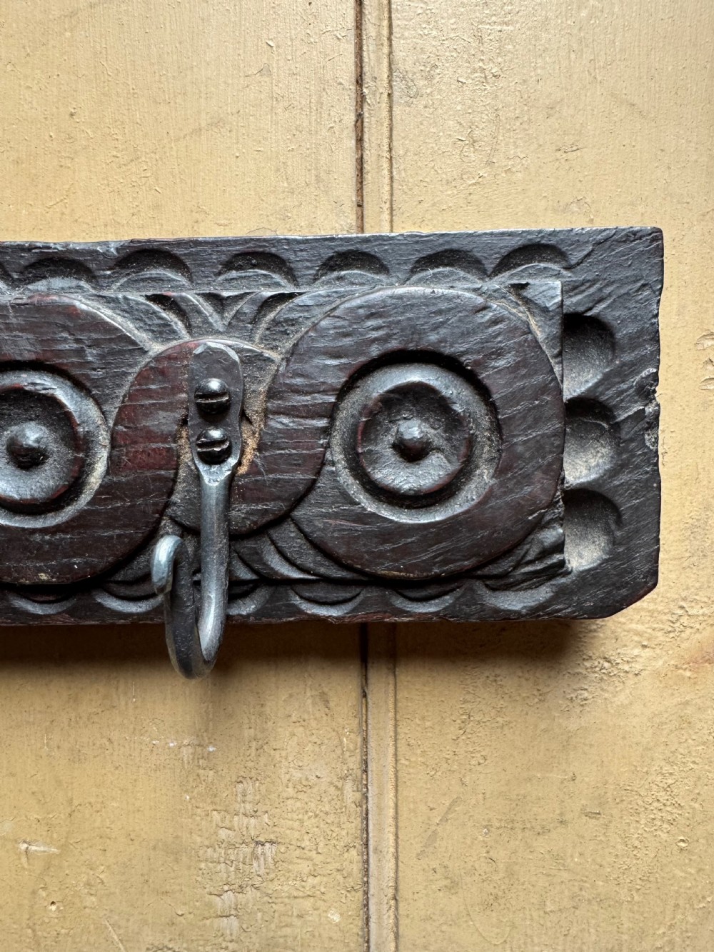 A dark carved oak wooden panel featuring relief carved circular scroll motifs and fitted with two dark metal hooks. View III