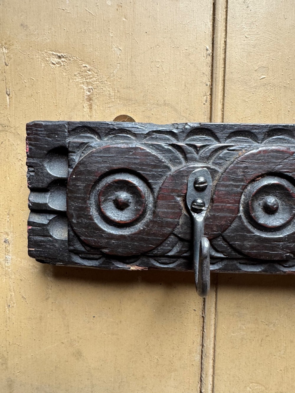 A dark carved oak wooden panel featuring relief carved circular scroll motifs and fitted with two dark metal hooks. View IV
