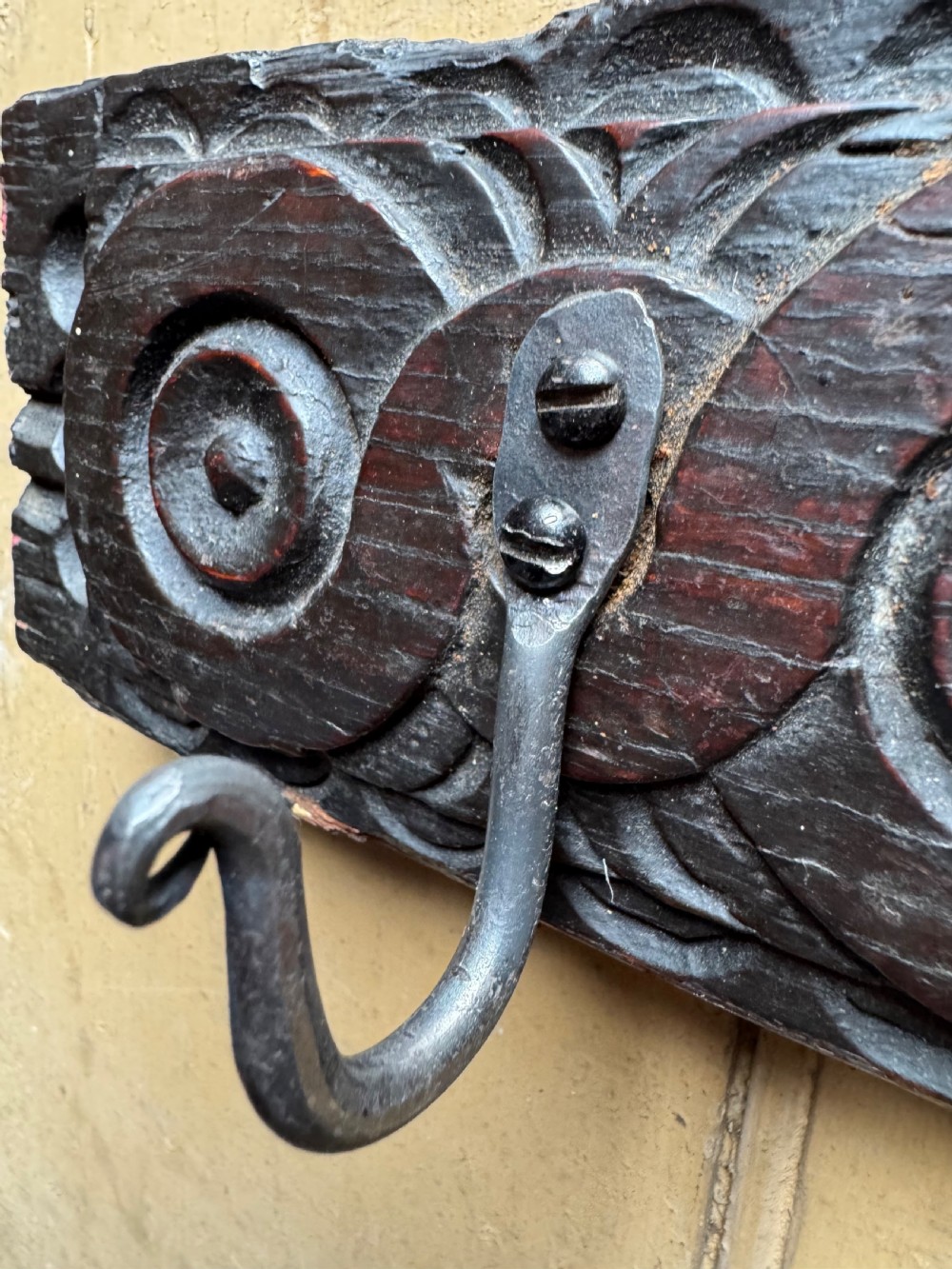 A dark carved oak wooden panel featuring relief carved circular scroll motifs and fitted with two dark metal hooks. View V