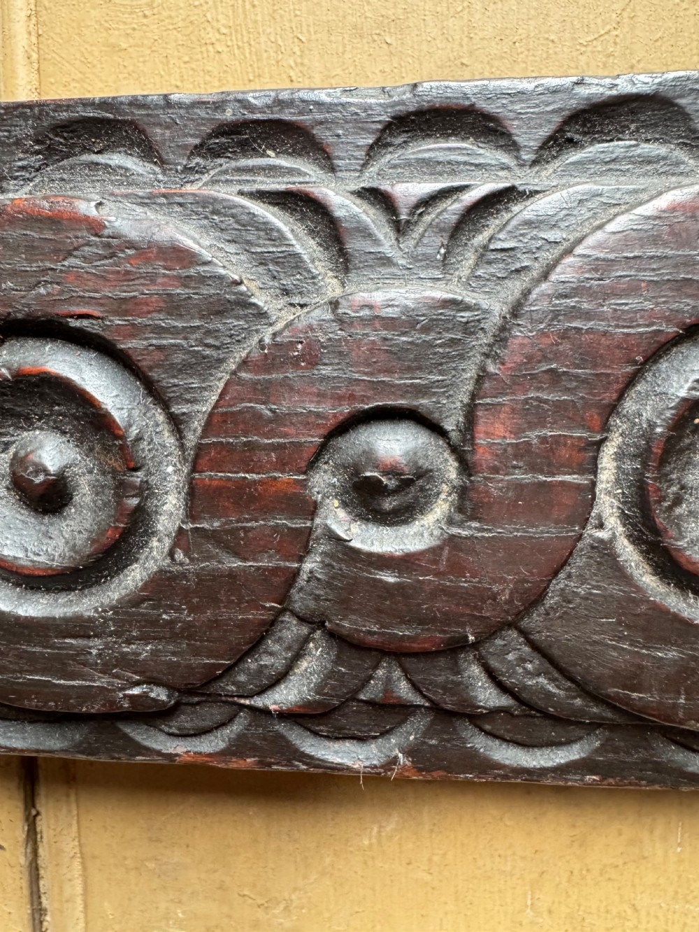 A dark carved oak wooden panel featuring relief carved circular scroll motifs and fitted with two dark metal hooks. View VII