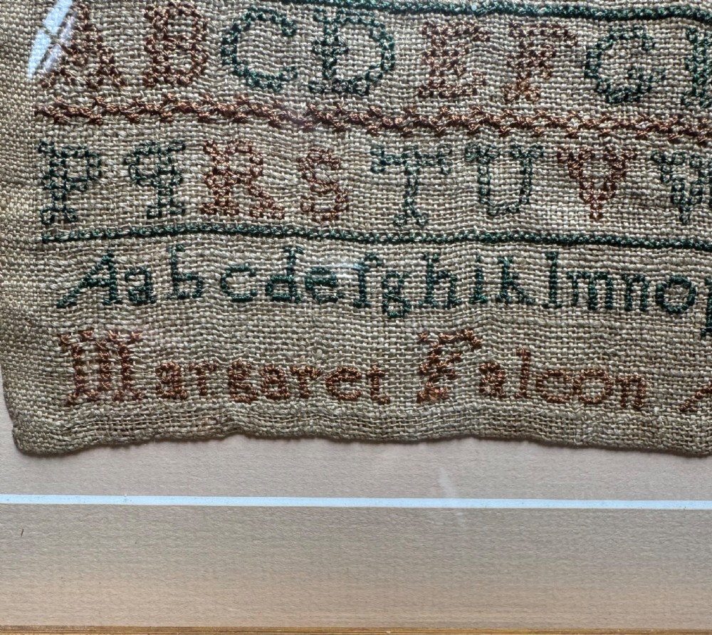 A framed nineteenth-century needlework sampler stitched with alphabet rows, numbers, and a signature on a natural linen ground within a gilded frame. View VI