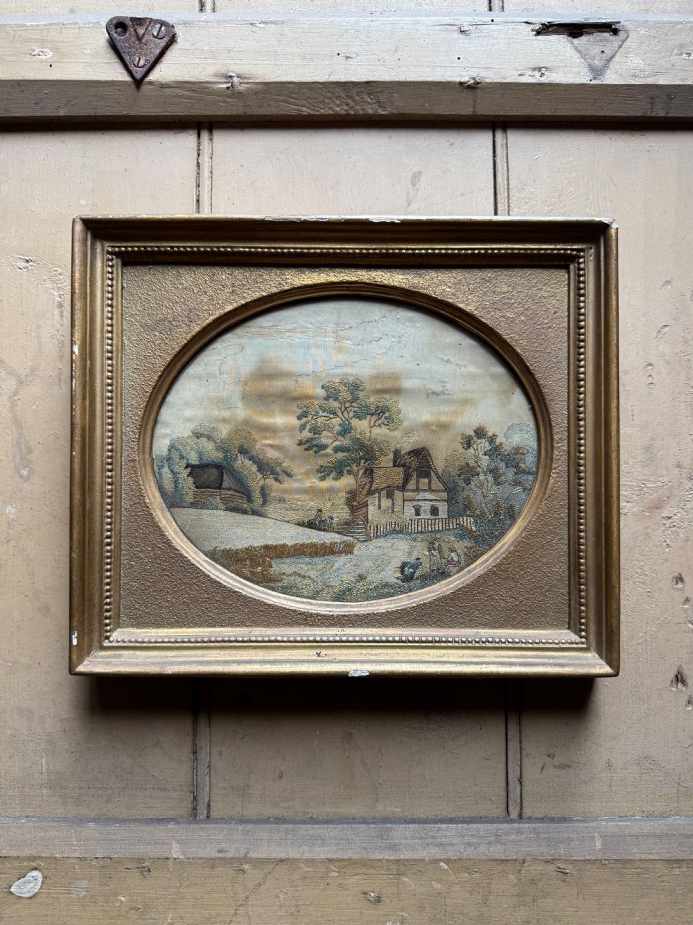 A framed rectangular gold gilded picture frame featuring an oval aperture surrounding a hand-embroidered silk landscape scene with a cottage and trees.