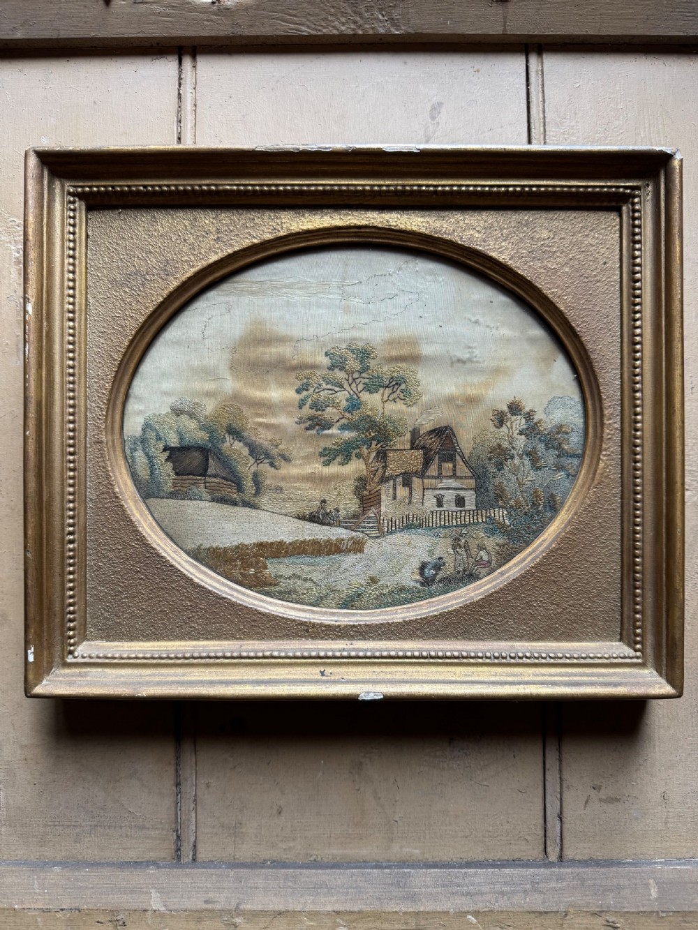 A framed rectangular gold gilded picture frame featuring an oval aperture surrounding a hand-embroidered silk landscape scene with a cottage and trees. View II