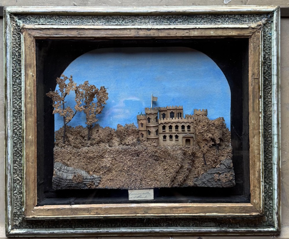 A framed nineteenth-century three-dimensional cork diorama depicting a castle on a hill against a painted light blue backdrop inside a glazed moulded frame. View II