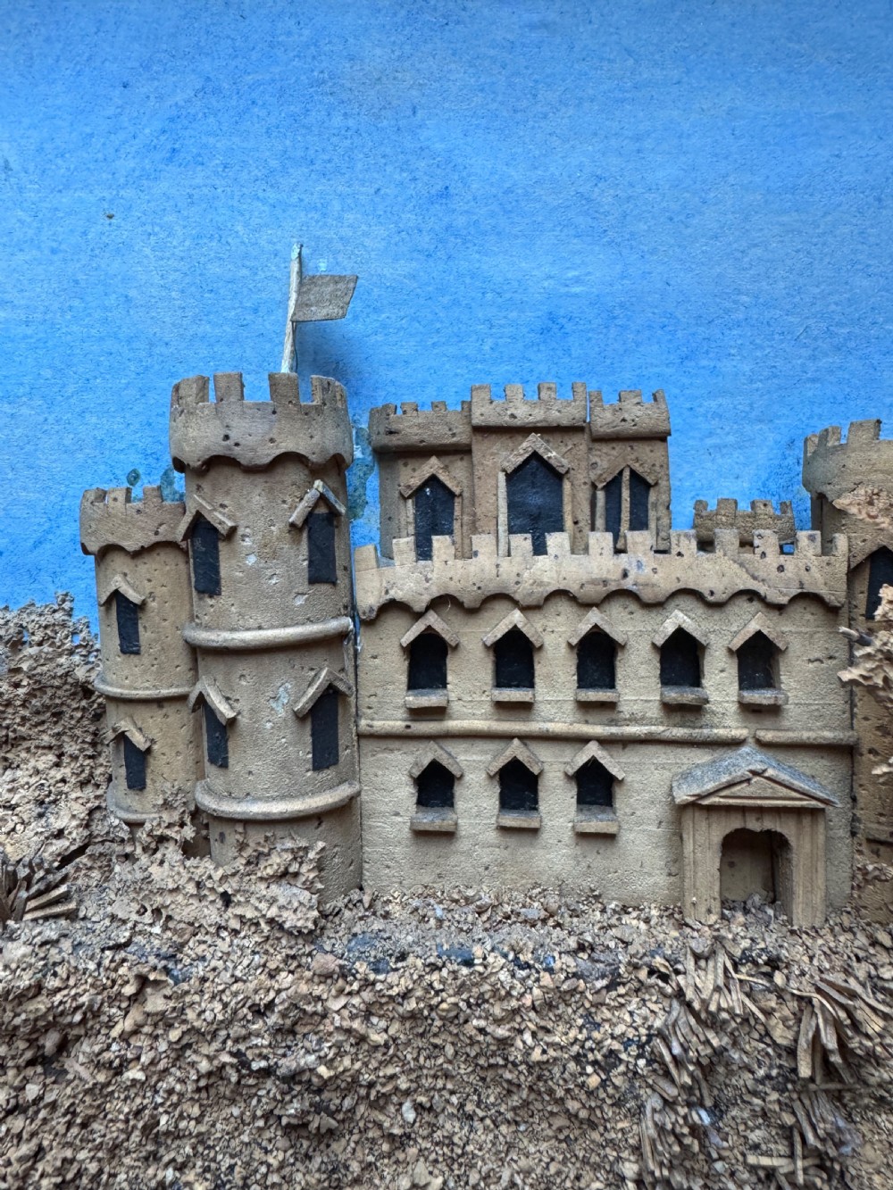 A framed nineteenth-century three-dimensional cork diorama depicting a castle on a hill against a painted light blue backdrop inside a glazed moulded frame. View XII
