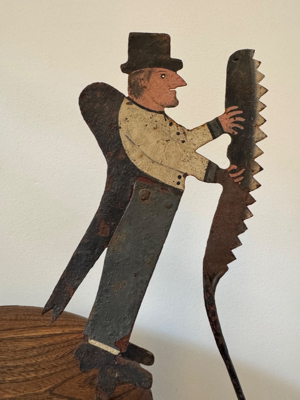A painted metal folk art figure depicting a sawyer wearing a top hat and holding a serrated saw with aged paint loss and oxidised patina.