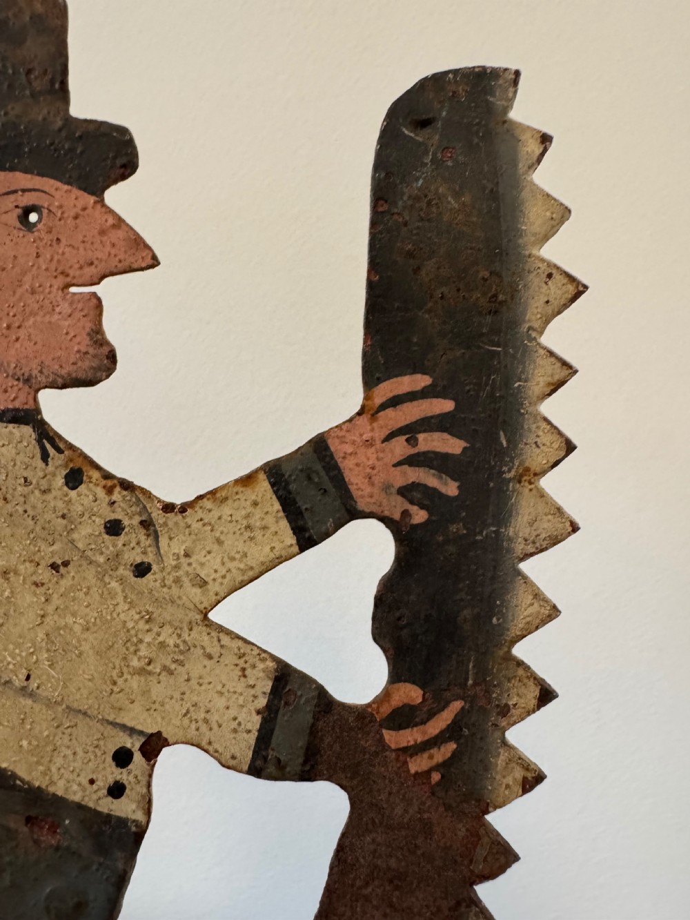 A painted metal folk art figure depicting a sawyer wearing a top hat and holding a serrated saw with aged paint loss and oxidised patina. View III