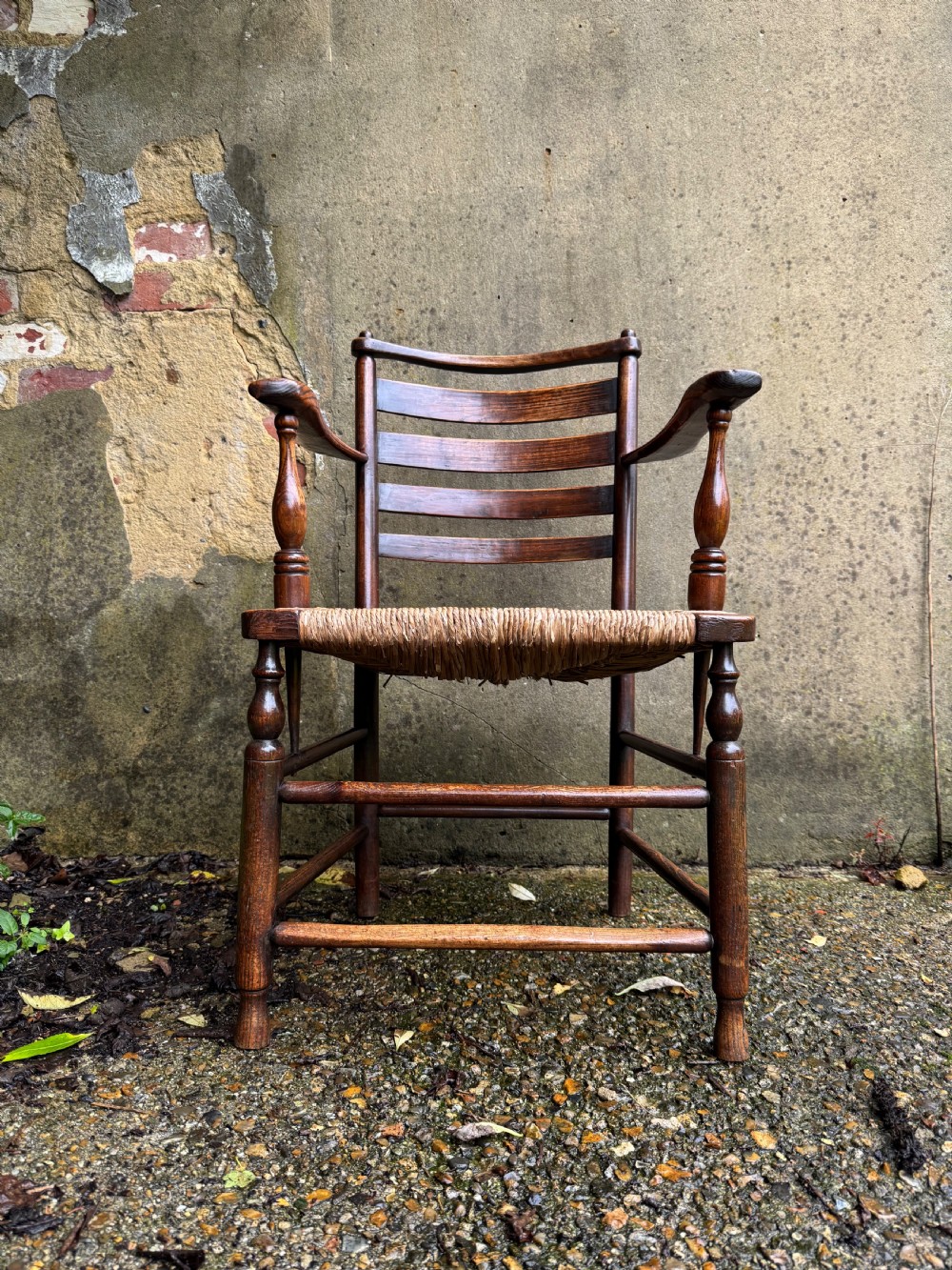 An antique dark wood Sussex armchair featuring a slatted backrest, turned front legs and stretchers, shaped armrests, and a hand-woven rush seat.