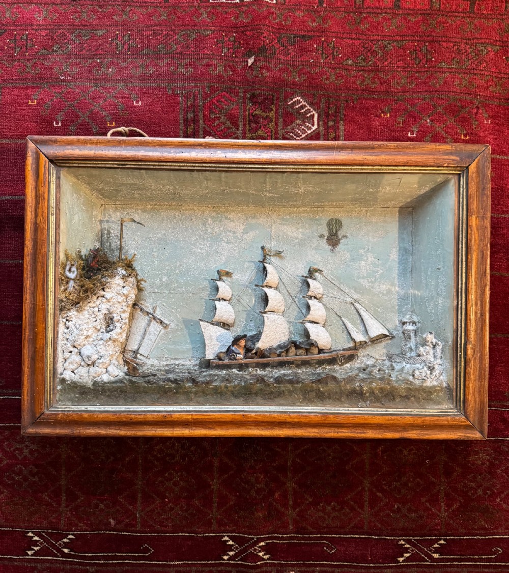 A framed shadow box diorama depicting a three-masted sailing ship on a painted plaster sea beside a rocky cliff and lighthouse.