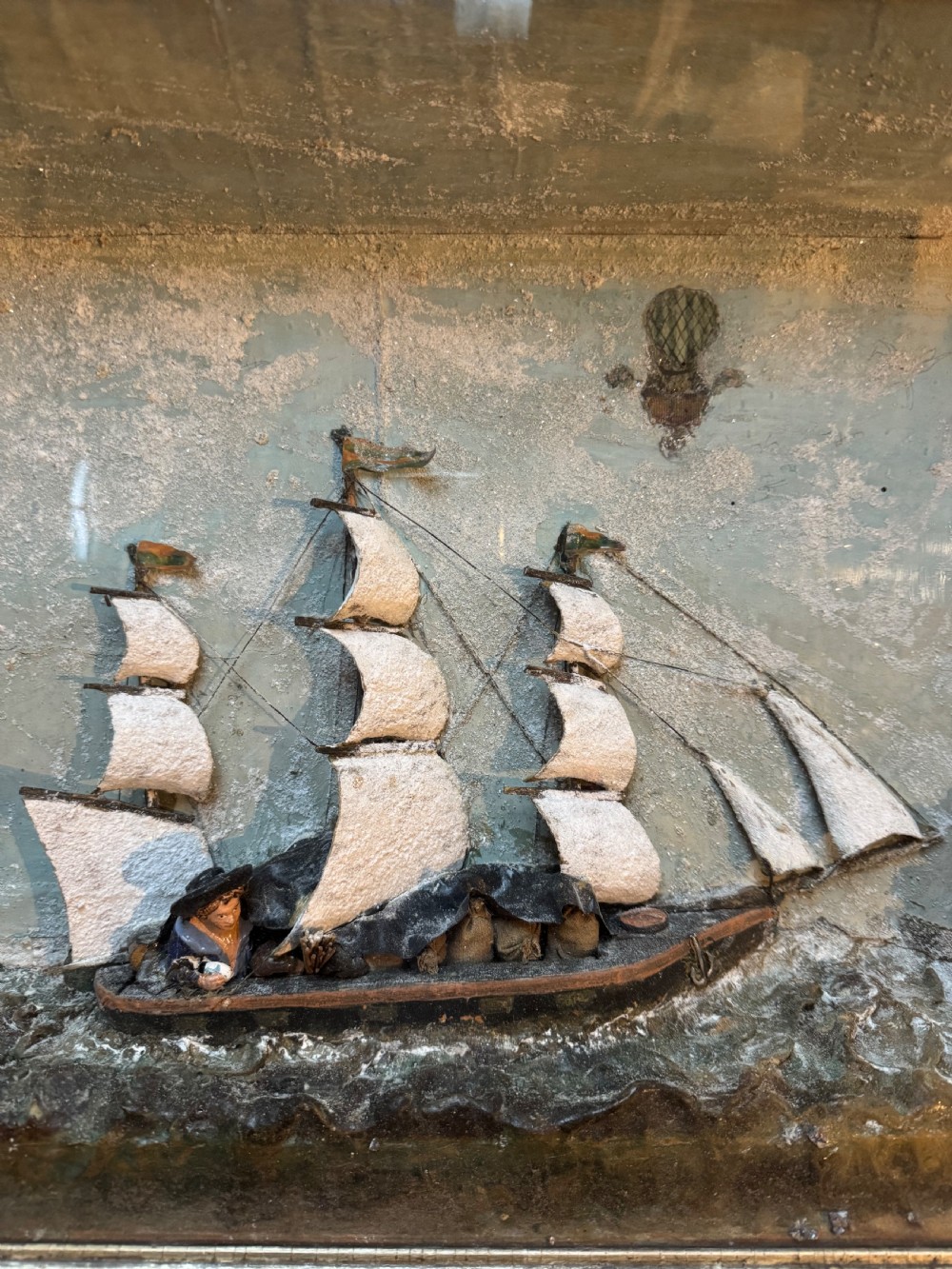 A framed shadow box diorama depicting a three-masted sailing ship on a painted plaster sea beside a rocky cliff and lighthouse. View III
