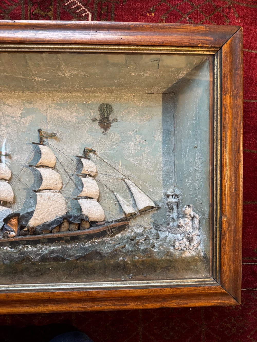 A framed shadow box diorama depicting a three-masted sailing ship on a painted plaster sea beside a rocky cliff and lighthouse. View IV
