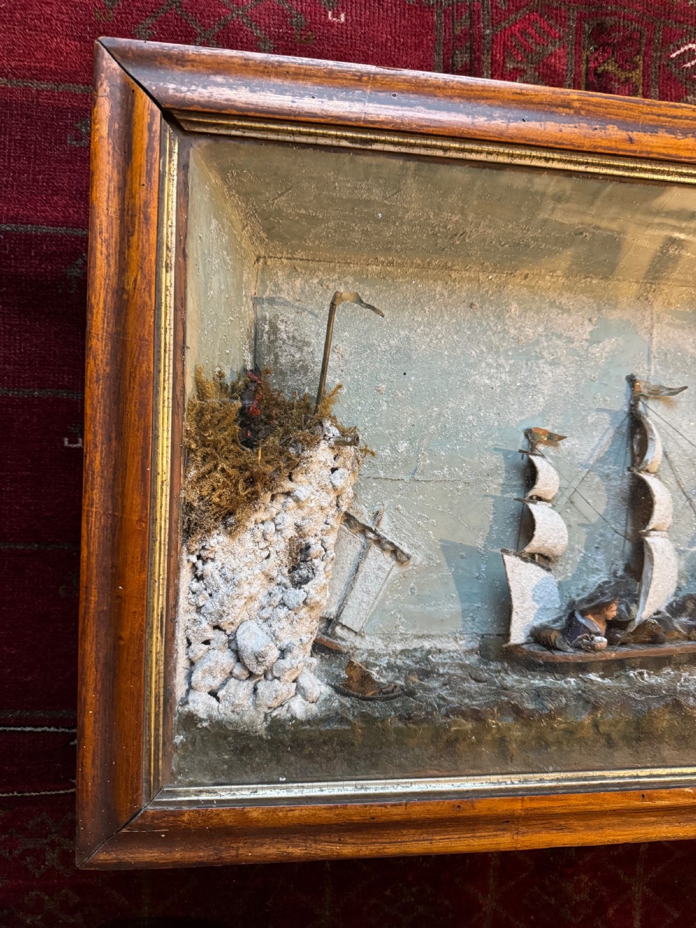 A framed shadow box diorama depicting a three-masted sailing ship on a painted plaster sea beside a rocky cliff and lighthouse. View V