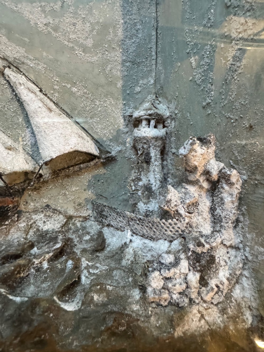 A framed shadow box diorama depicting a three-masted sailing ship on a painted plaster sea beside a rocky cliff and lighthouse. View VII