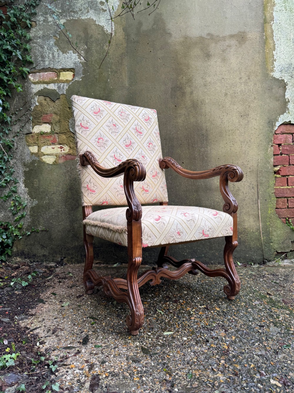 A French style carved walnut armchair featuring a tall backrest, scrolled armrests, patterned cream and pink upholstery, and intricately carved legs.