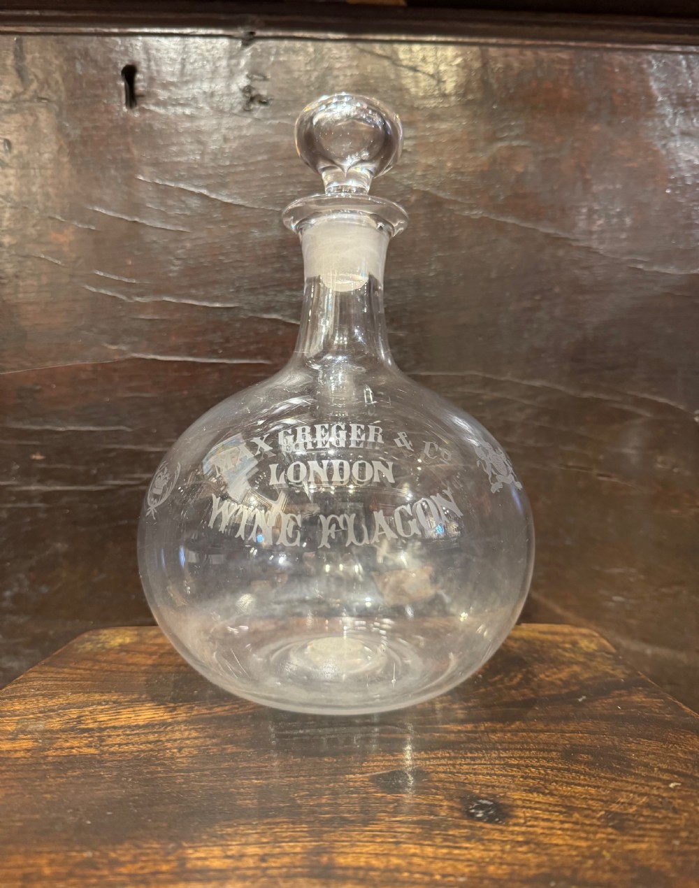 A clear bulbous glass wine flagon featuring etched lettering reading Max Greger and Co London Wine Flagon with a matching glass stopper.