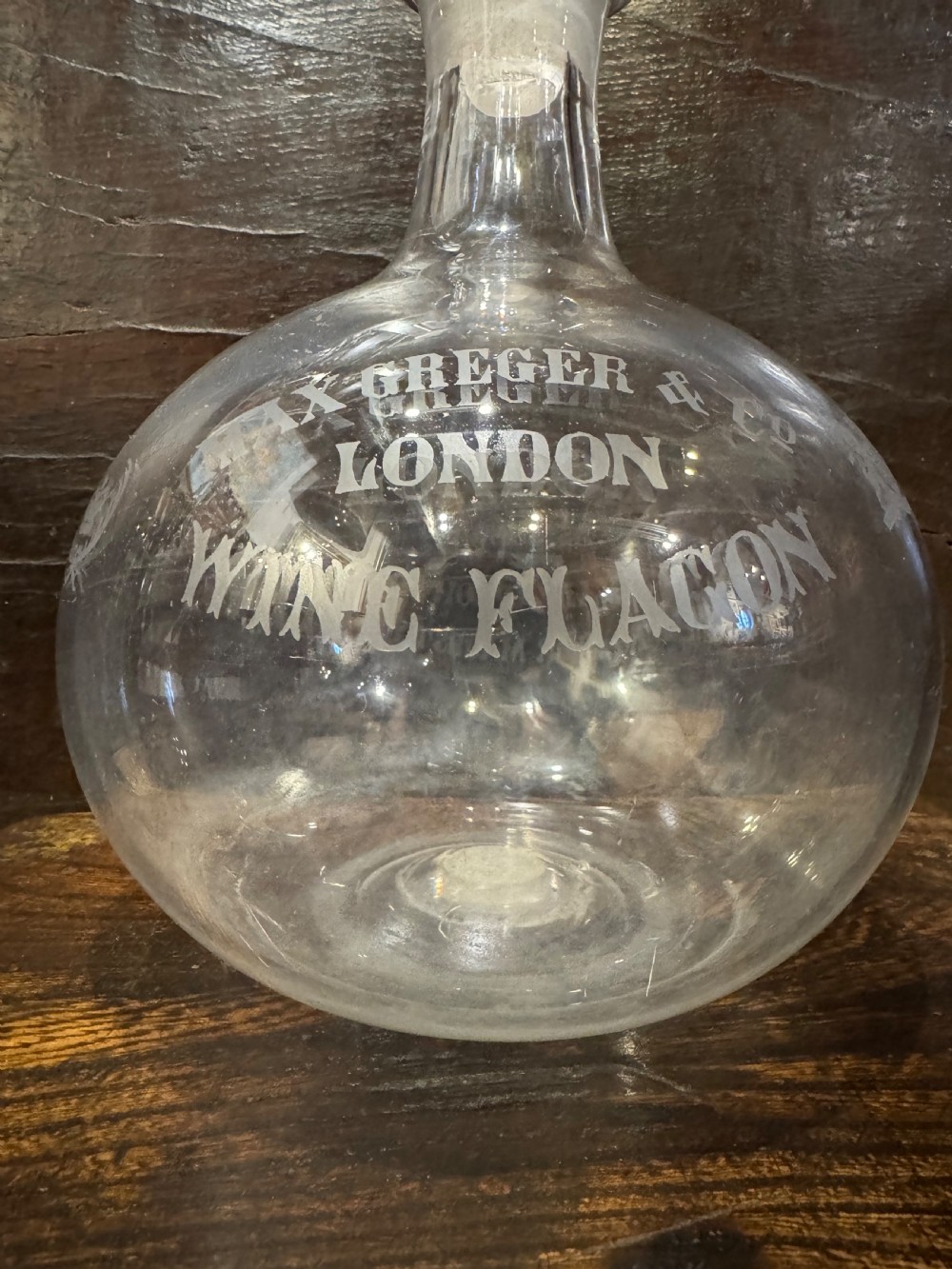 A clear bulbous glass wine flagon featuring etched lettering reading Max Greger and Co London Wine Flagon with a matching glass stopper. View II