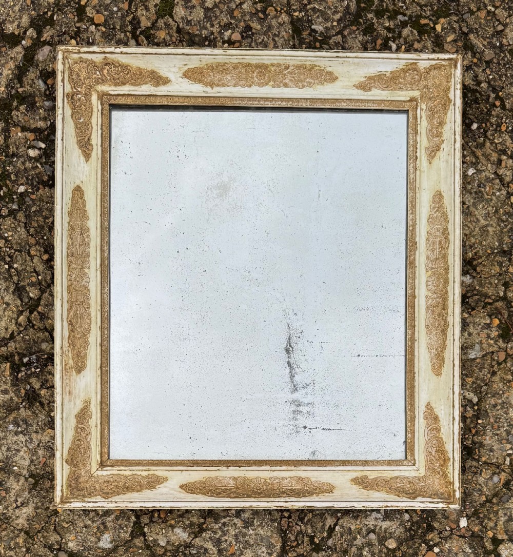 A rectangular antique wall mirror featuring a distressed cream-painted timber frame decorated with gilded corner spandrels and foxed mirror glass.
