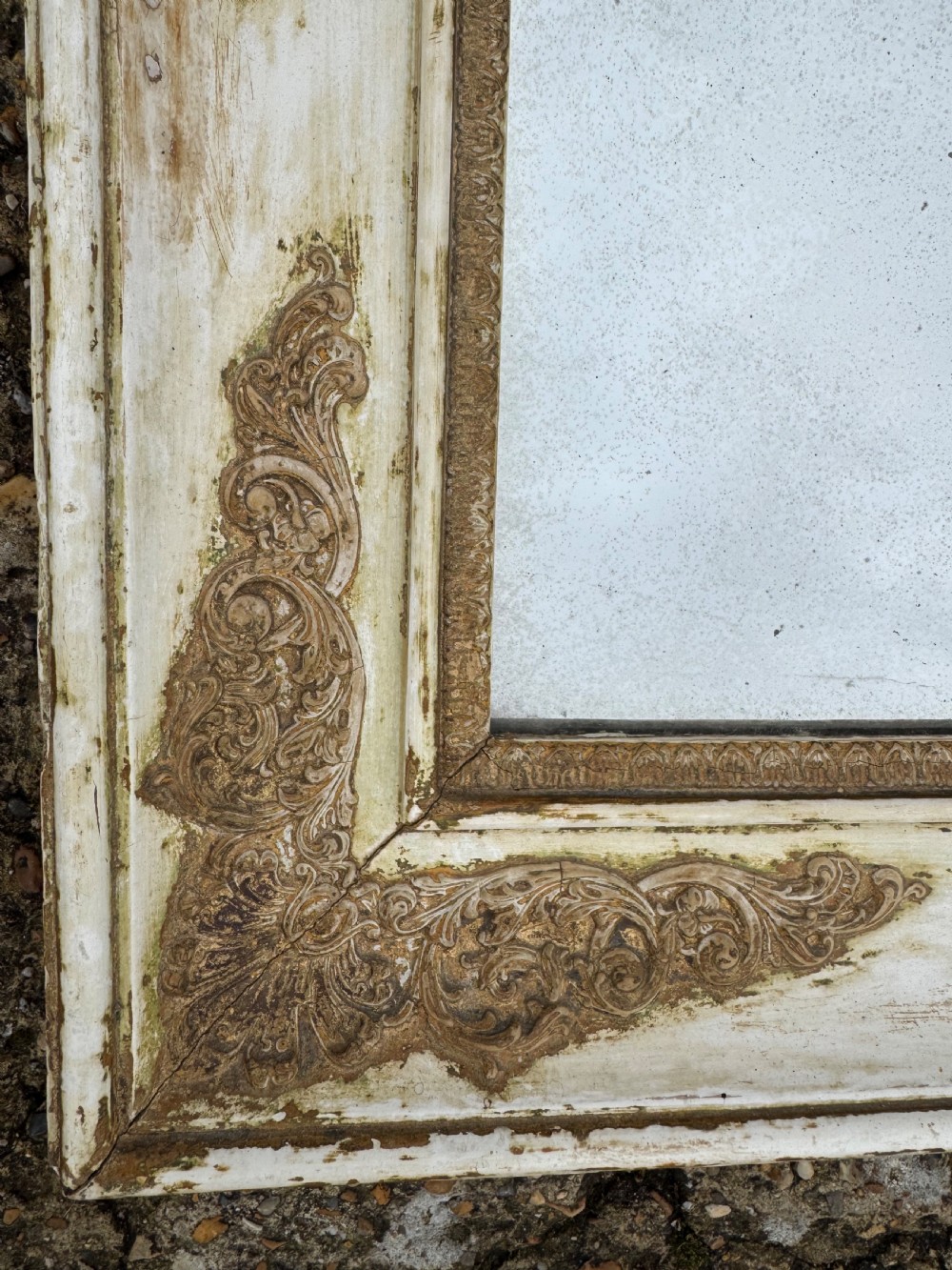 A rectangular antique wall mirror featuring a distressed cream-painted timber frame decorated with gilded corner spandrels and foxed mirror glass. View V