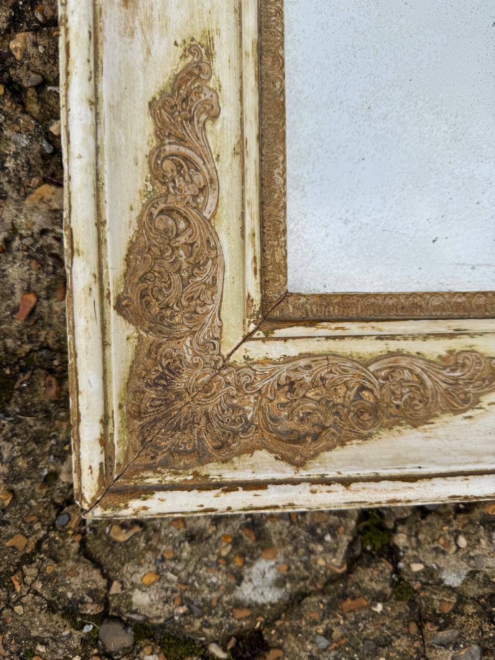 A rectangular antique wall mirror featuring a distressed cream-painted timber frame decorated with gilded corner spandrels and foxed mirror glass. View VI