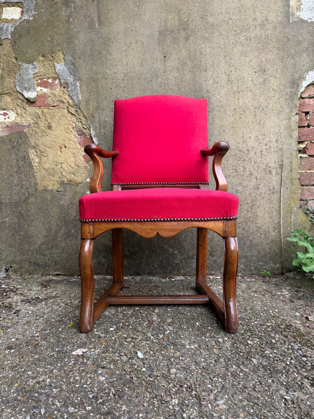 An antique French wooden armchair featuring red fabric upholstery, scrolled arms, brass stud trimming, and a rectangular stretcher base.