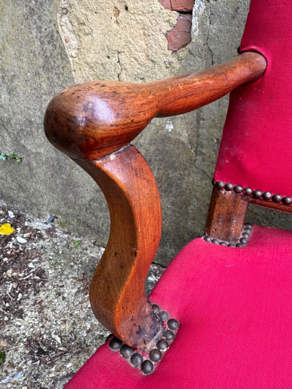 An antique French wooden armchair featuring red fabric upholstery, scrolled arms, brass stud trimming, and a rectangular stretcher base. View V