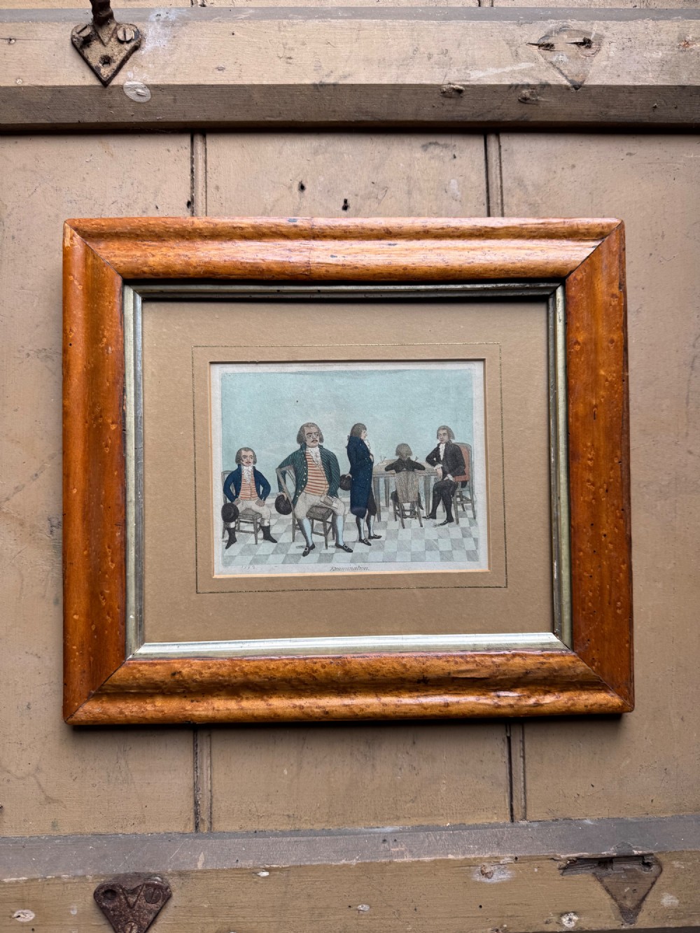 A framed Georgian hand-coloured etching titled Examination depicting figures in 18th-century dress set within a thick, light brown wood frame.