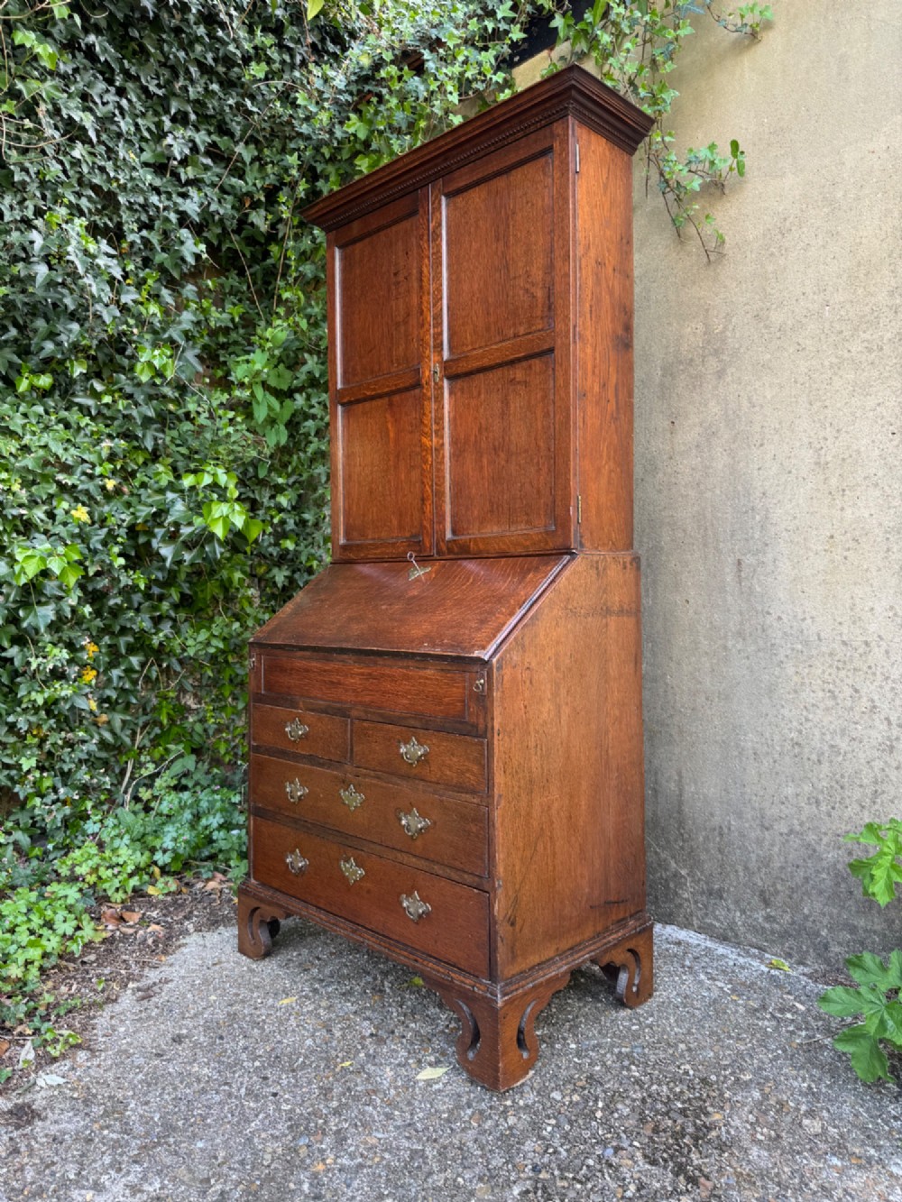 An antique warm brown oak bureau bookcase featuring twin fielded panel upper doors, a slant-front desk, and multiple drawers with brass hardware.
