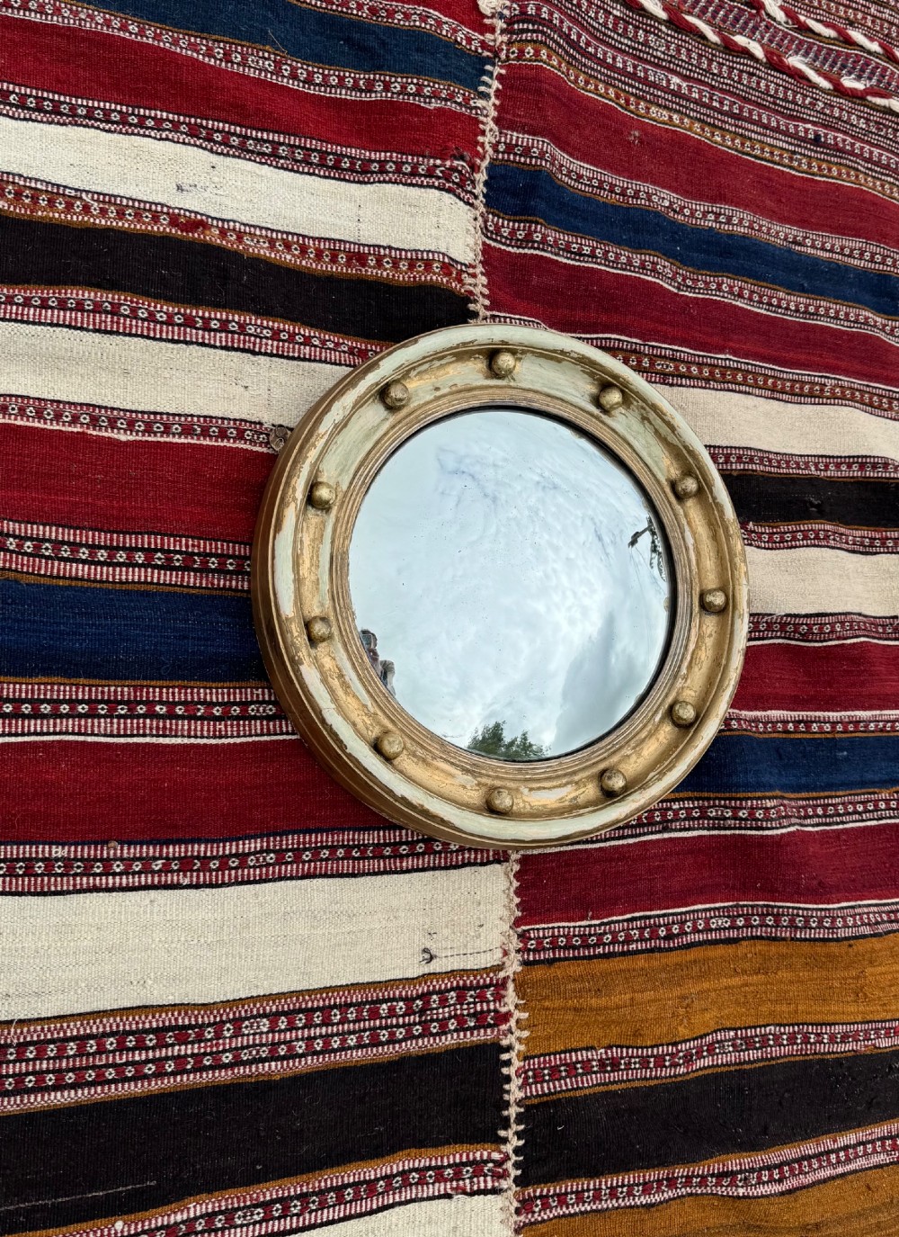 A circular antique convex mirror featuring a gilded and painted frame decorated with spherical balls along the inner rim.