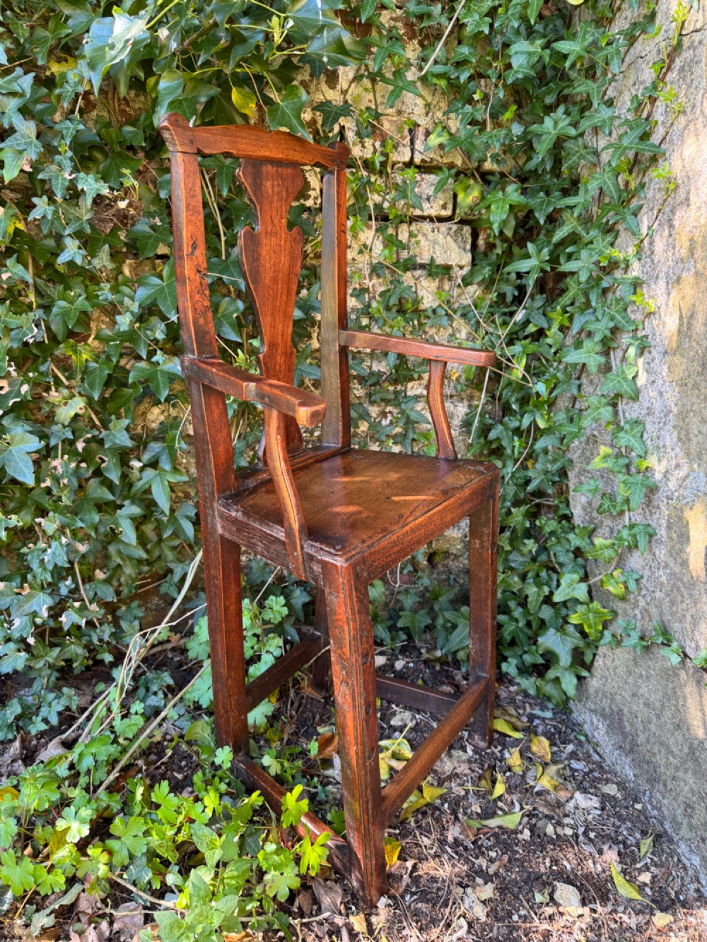 A dark brown Georgian wooden high chair with a central splat back, shaped armrests, square legs, and low wooden stretchers.