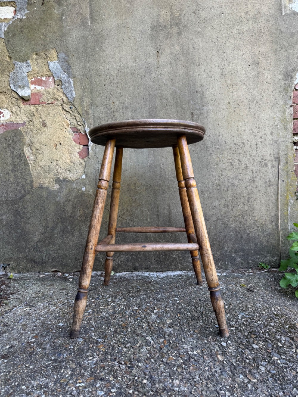 A rustic circular wooden stool featuring four splayed turned legs supported by horizontal timber stretchers.