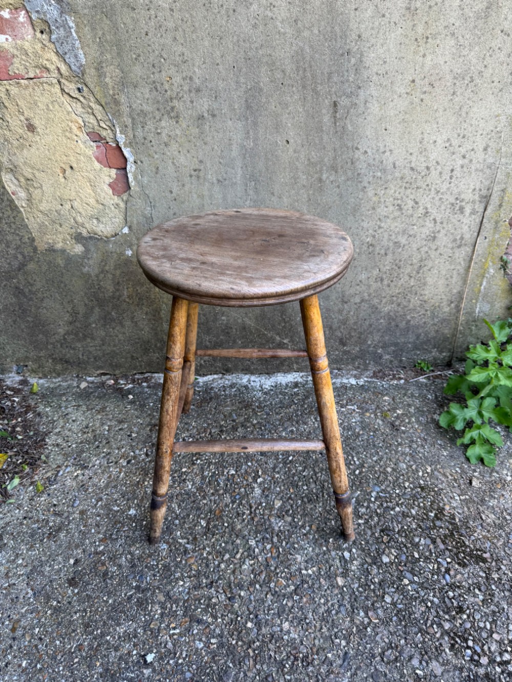 A rustic circular wooden stool featuring four splayed turned legs supported by horizontal timber stretchers. View III