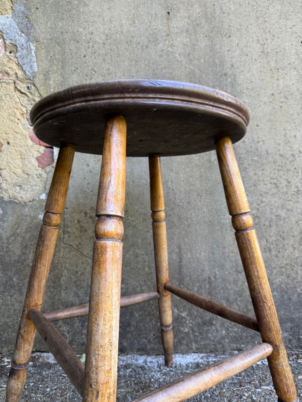 A rustic circular wooden stool featuring four splayed turned legs supported by horizontal timber stretchers. View VIII