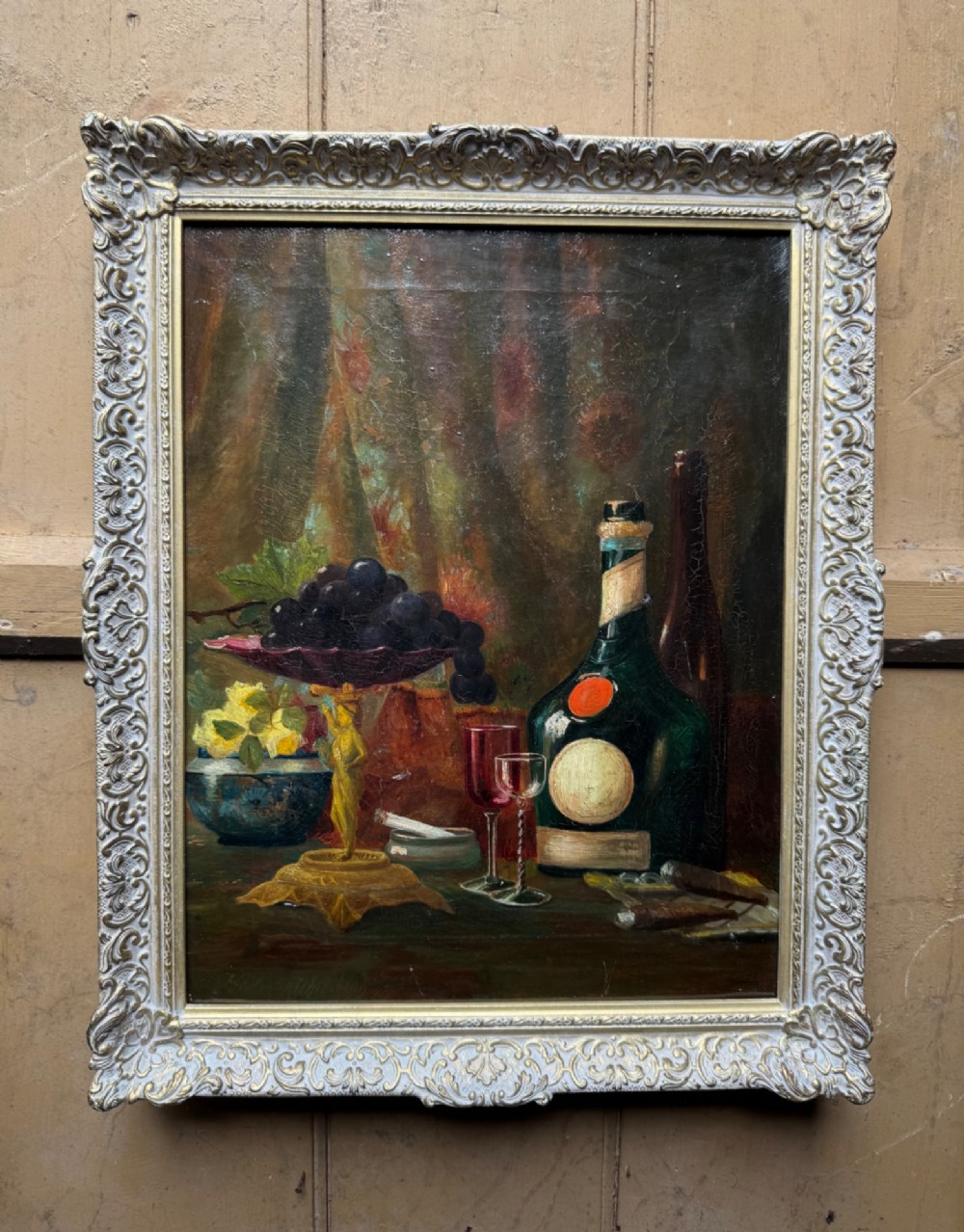 Antique framed oil painting depicting a still life composition with fruit, glassware, and bottles set in an ornate swept frame.