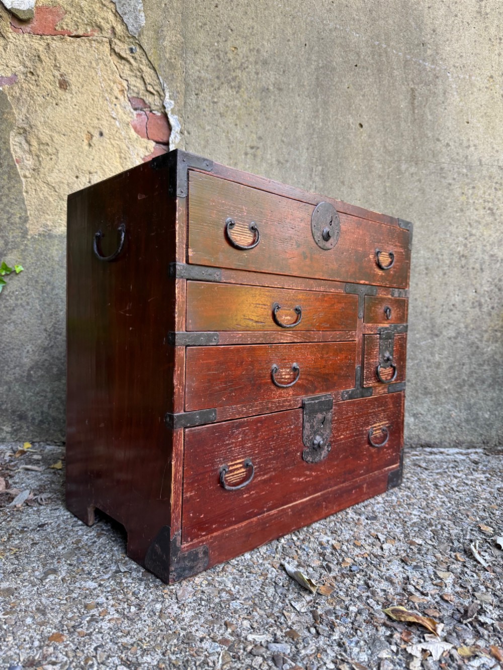 A reddish brown lacquered Japanese wooden tansu chest featuring iron corner plates, circular lock fittings, side carrying handles, and multiple graduated drawers.