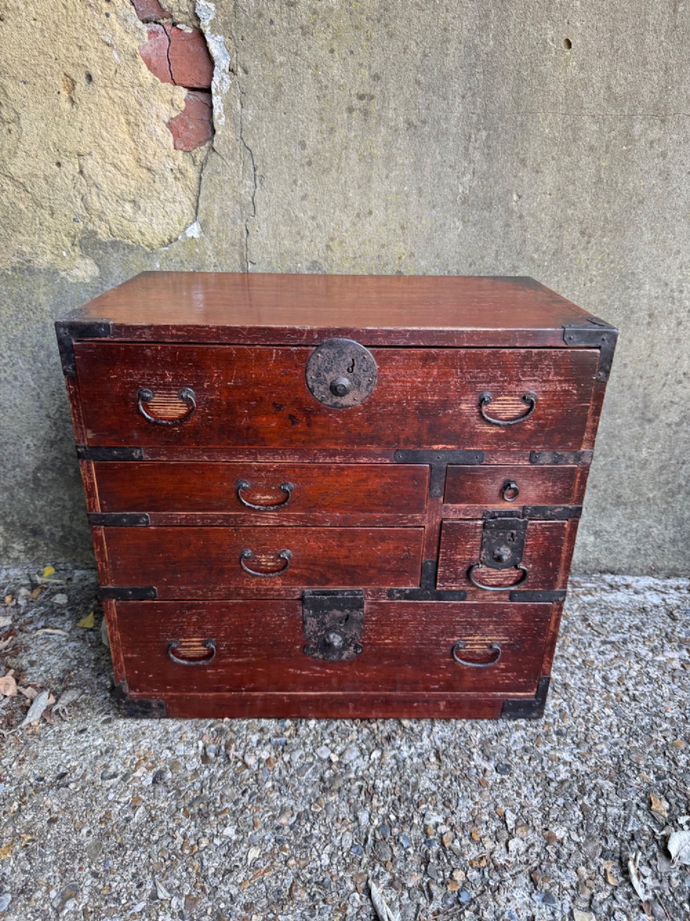 A reddish brown lacquered Japanese wooden tansu chest featuring iron corner plates, circular lock fittings, side carrying handles, and multiple graduated drawers. View III