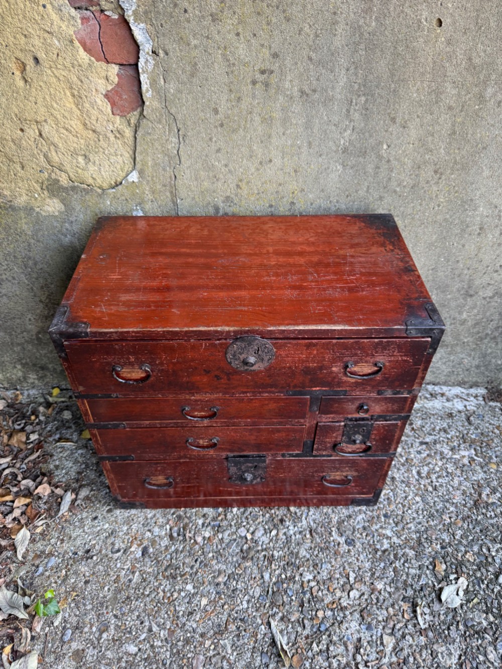 A reddish brown lacquered Japanese wooden tansu chest featuring iron corner plates, circular lock fittings, side carrying handles, and multiple graduated drawers. View IV