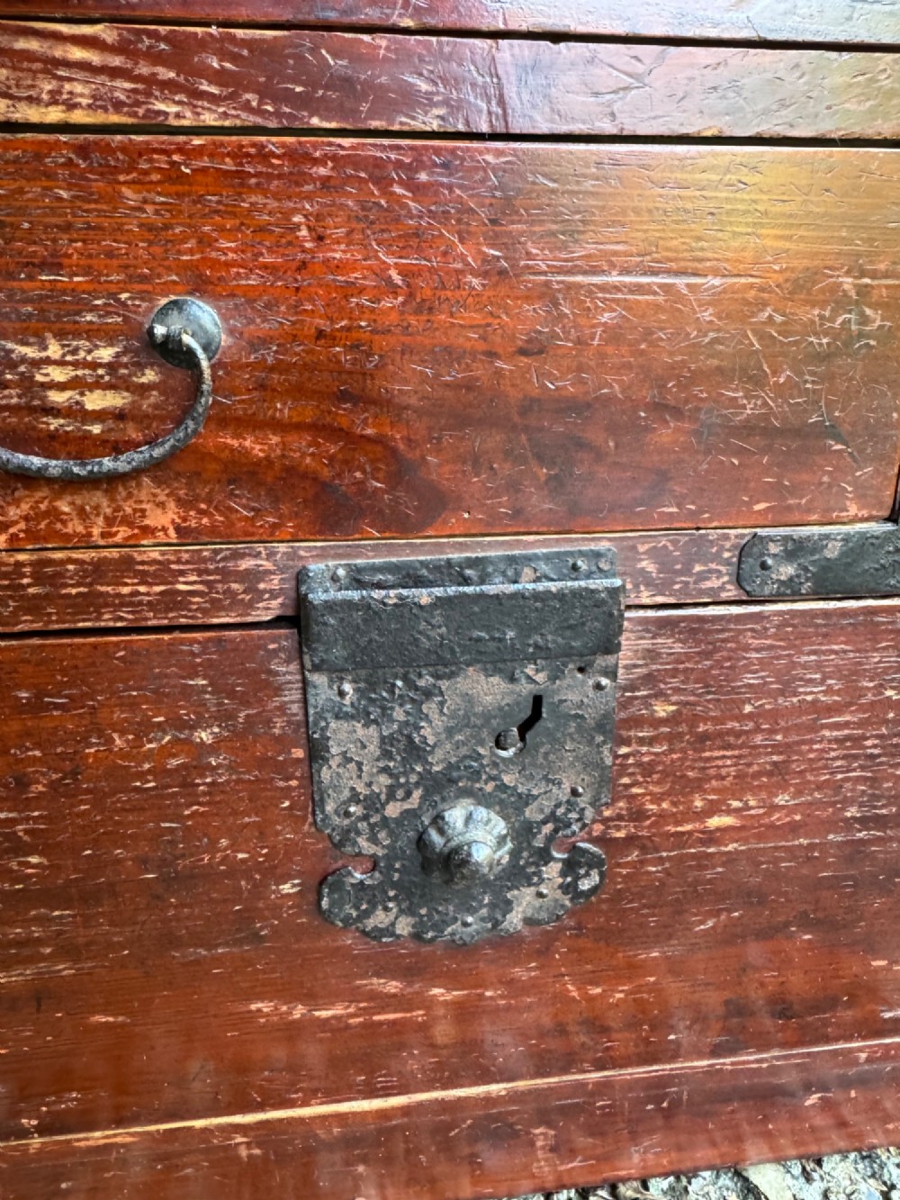 A reddish brown lacquered Japanese wooden tansu chest featuring iron corner plates, circular lock fittings, side carrying handles, and multiple graduated drawers. View VI